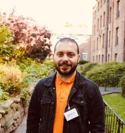 Profile photo of Archaeology PhD student Batuhan Özdemir. He wears an orange shirt and black denim jacket. In the background is one of the Durham University college spaces with various plants in the sunshine..